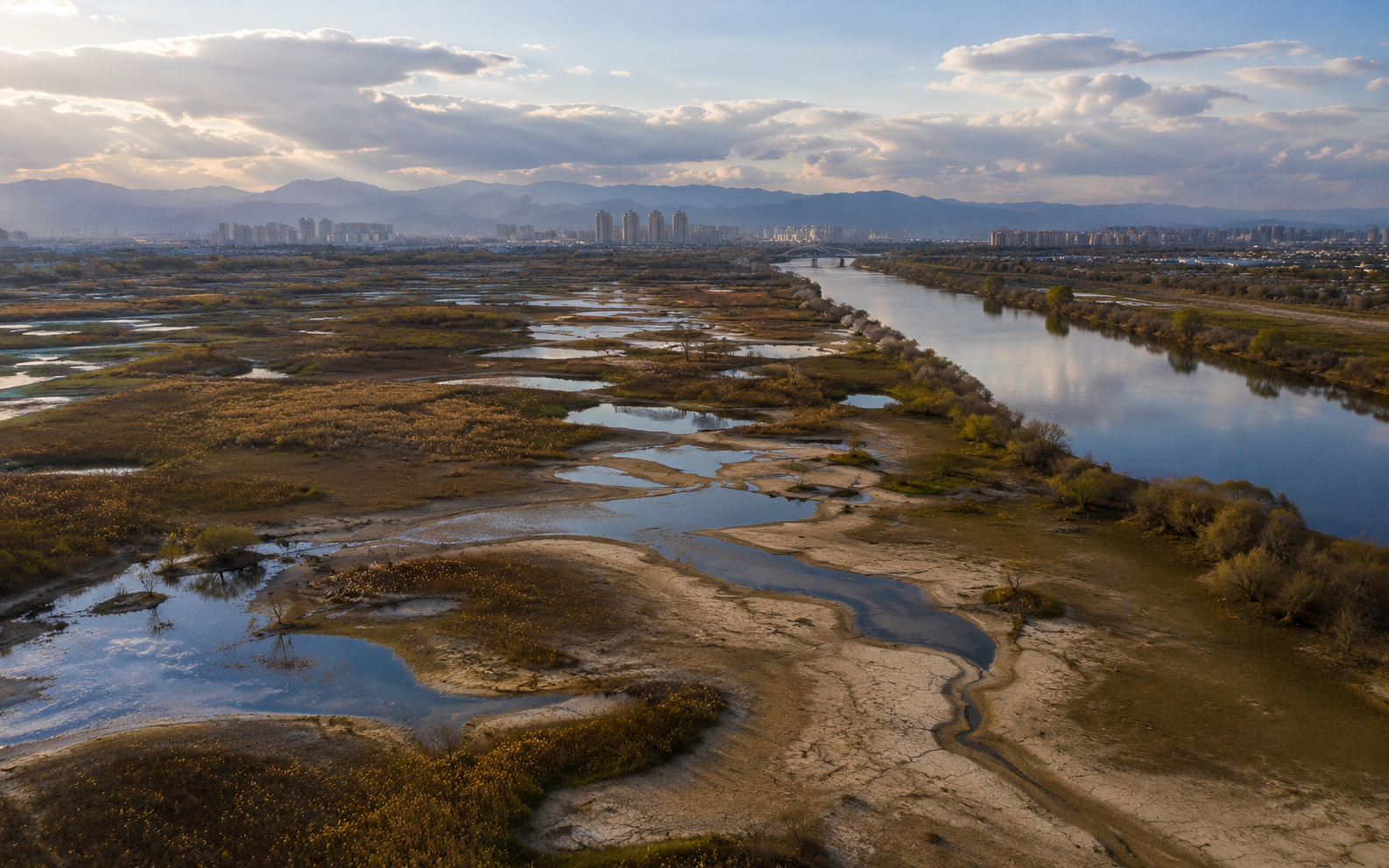 Dry wetland and river landscape near a distant city under changing light